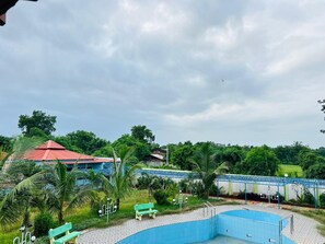 A swimming pool with a blue border, surrounded by greenery and a red-roofed building. - Vrundavan Village Resort and Home Stay (Garudeshwar)