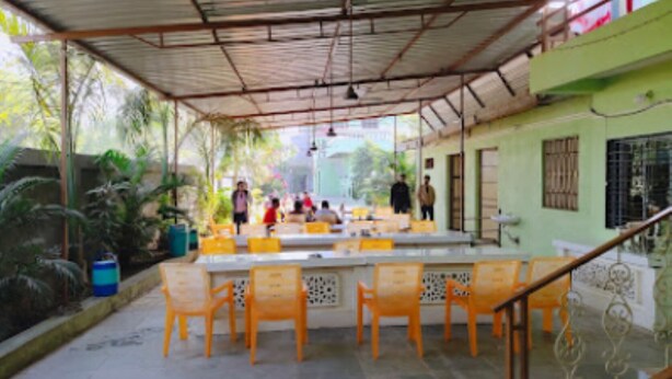 An outdoor covered seating area with tables and chairs, surrounded by greenery and a building with a green facade. - Vrundavan Village Resort and Home Stay (Garudeshwar)