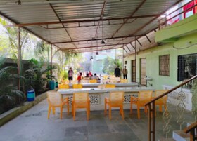 An outdoor covered seating area with tables and chairs, surrounded by greenery and a building with a green facade.