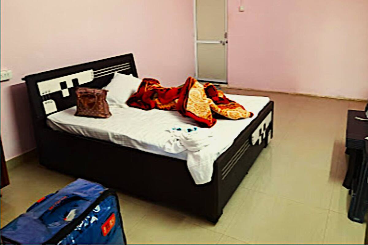 A single bed with a black frame, white bedding, and a red blanket. There is a blue bag on the floor beside the bed.