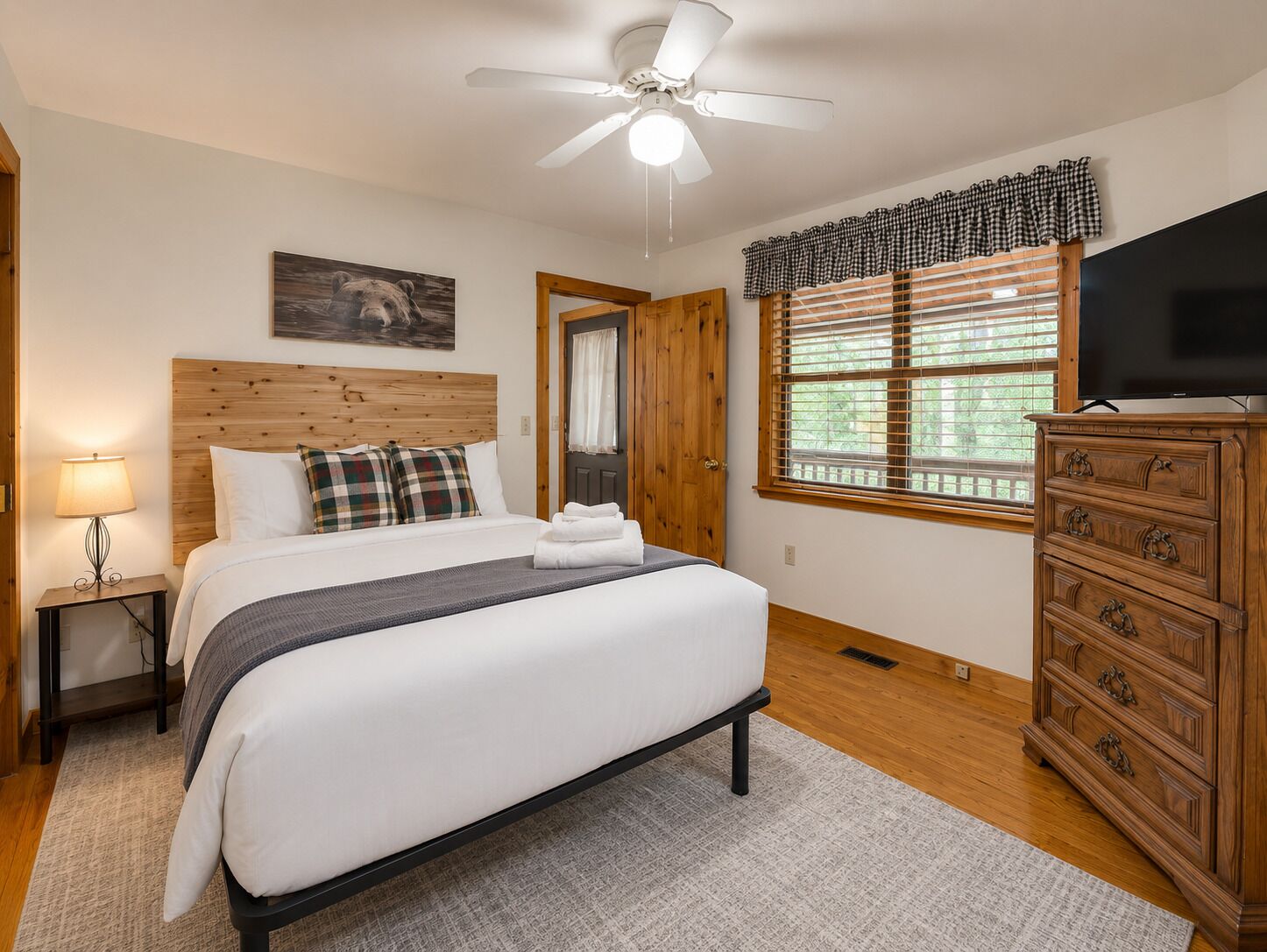 Une chambre à coucher comprenant un lit, une table de chevet, une commode, un ventilateur de plafond, une fenêtre avec des stores et un tableau accroché au mur.