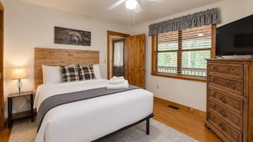 Une chambre à coucher comprenant un lit, une table de chevet, une commode, un ventilateur de plafond, une fenêtre avec des stores et un tableau accroché au mur.