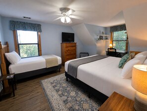 Une chambre avec deux lits, un ventilateur de plafond, une télévision, une commode, un bureau avec une lampe et une fenêtre avec des stores.