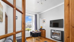 Una sala moderna con un sofá, una mesa de comedor con sillas, una chimenea, un televisor montado en la pared y un ventanal con vistas al exterior.