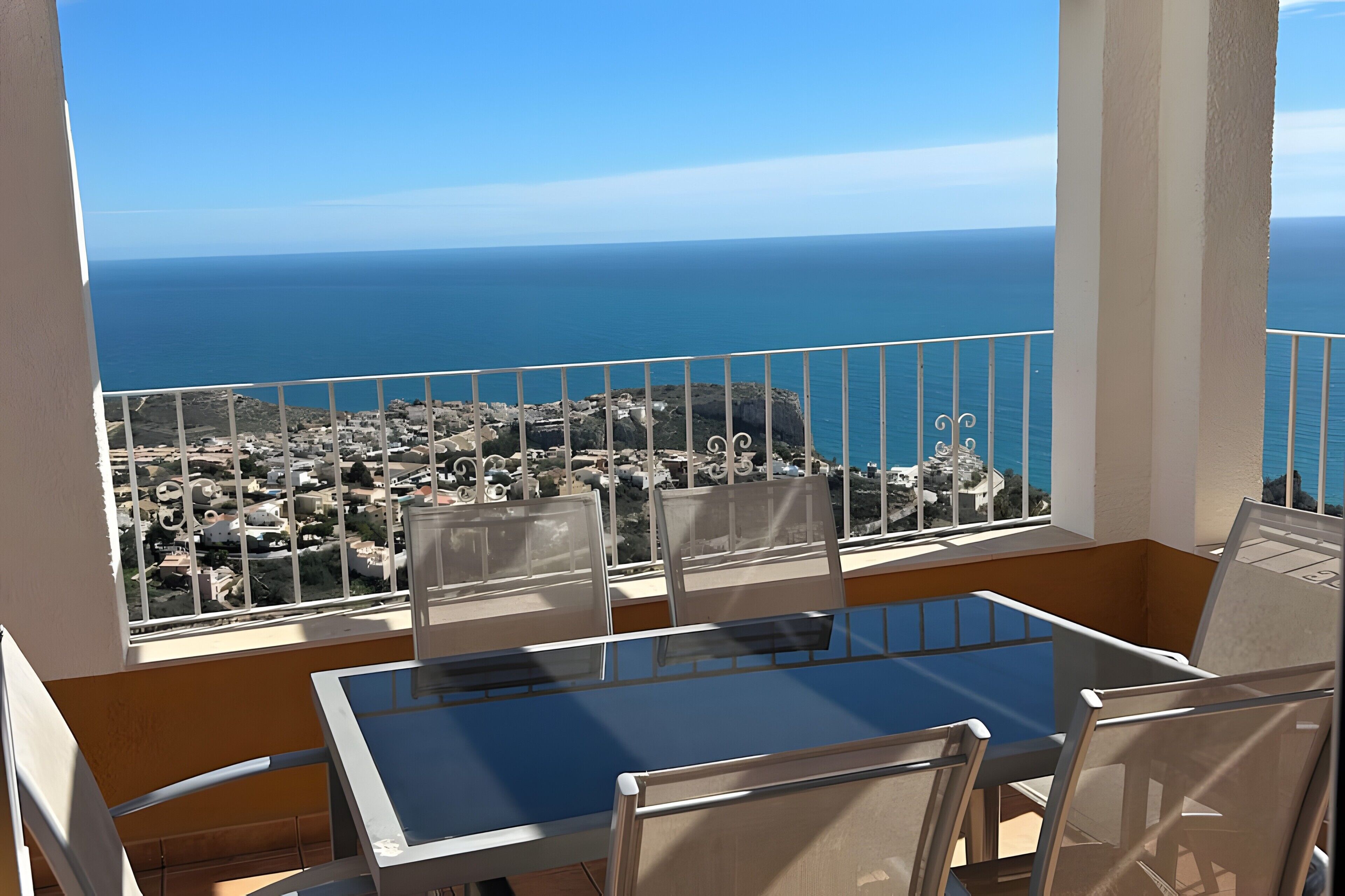 A balcony with a glass table and chairs, offering a view of the sea and a coastal town.