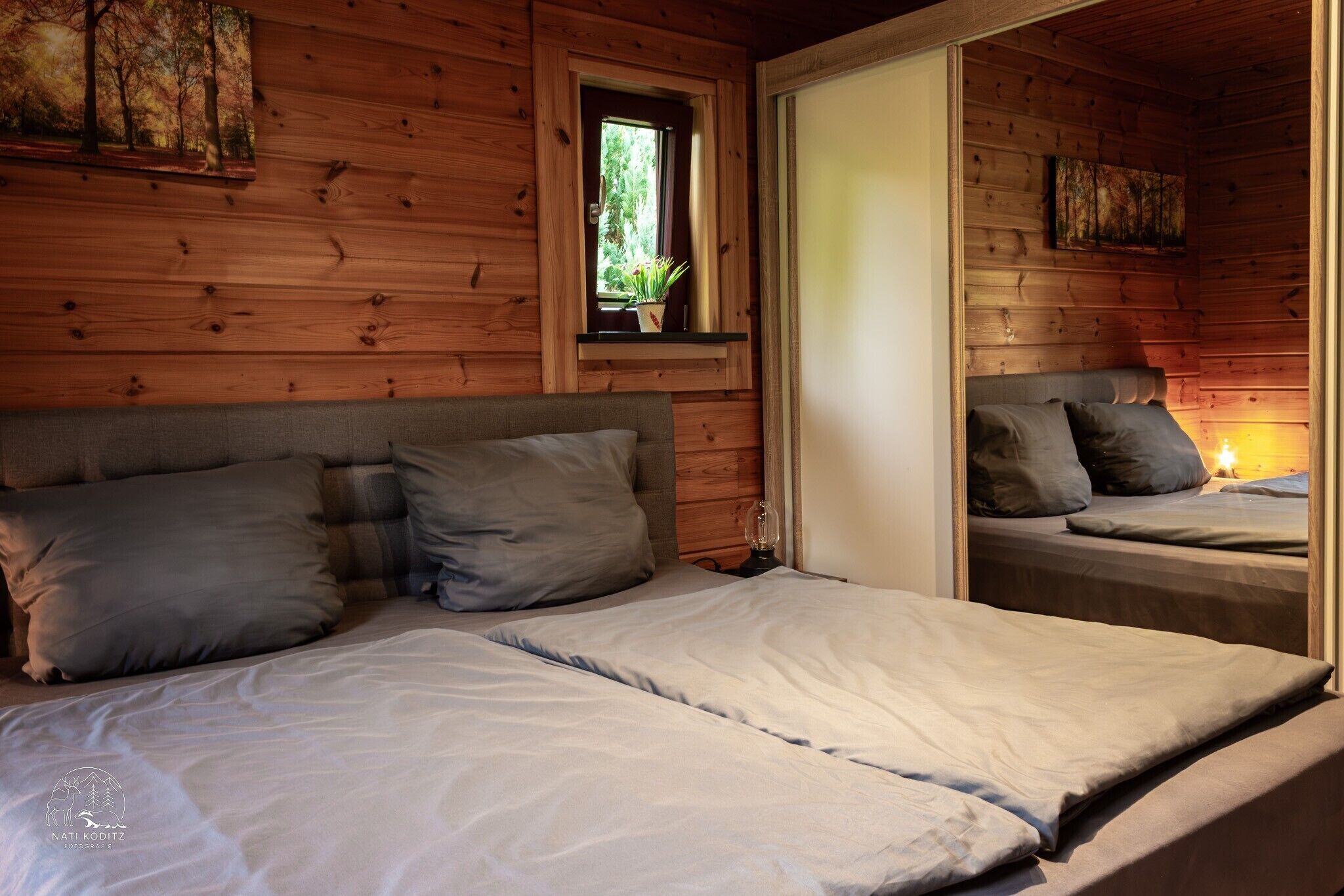 Une cabane en bois avec deux lits, une fenêtre ornée d’une plante et un miroir qui reflète la pièce.