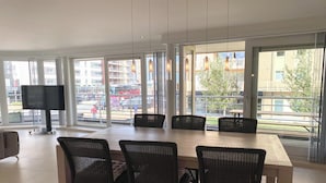 Un comedor amplio con una mesa grande y sillas de mimbre, ventanas grandes que ofrecen vistas de la calle y edificios cercanos.