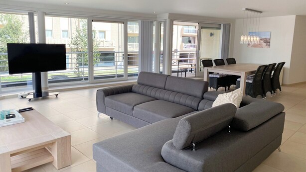 Una sala de estar moderna con un sofá seccional gris, un televisor de pantalla plana y un comedor con una mesa y sillas.
