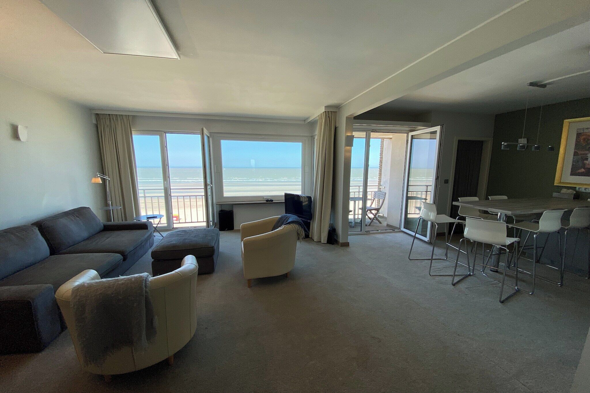 A spacious living area with a sofa, armchairs, and a dining table with chairs. Large windows offer a view of the beach.