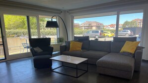 A modern living room with a grey sofa, a black armchair, and a coffee table. Large windows offer a view of the street and buildings outside. - Apartment in Nieuwpoort near Sandy Beaches (NIEUWPOORT)