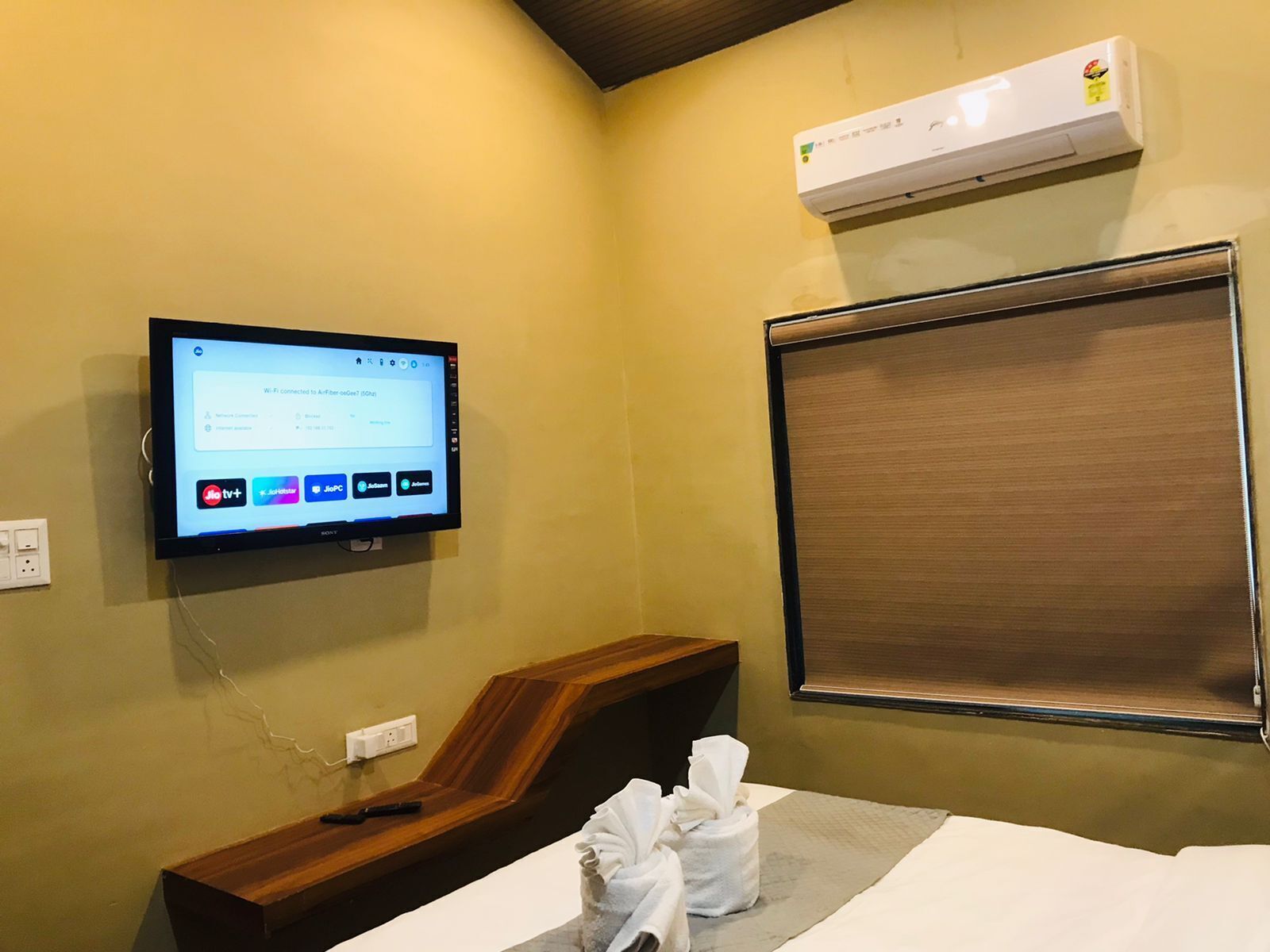 Een hotelkamer met een televisie waarop een scherm met een Wi-Fi verbinding wordt getoond, een houten nachtkastje, een raam met een zonwering en een wandairconditioner.