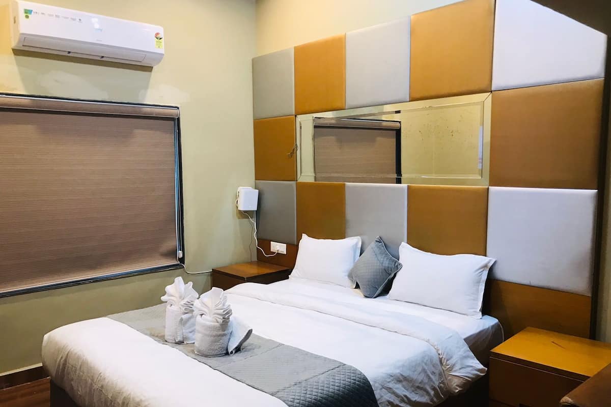 A hotel room with a bed, a bedside table, a nightstand, a wall-mounted air conditioner, and a window with blinds.