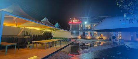 Uma área de refeições ao ar livre com mesas e cadeiras, um espaço de estar coberto e um edifício com uma placa que diz "KOSHI AERO CLUB & RESORT".