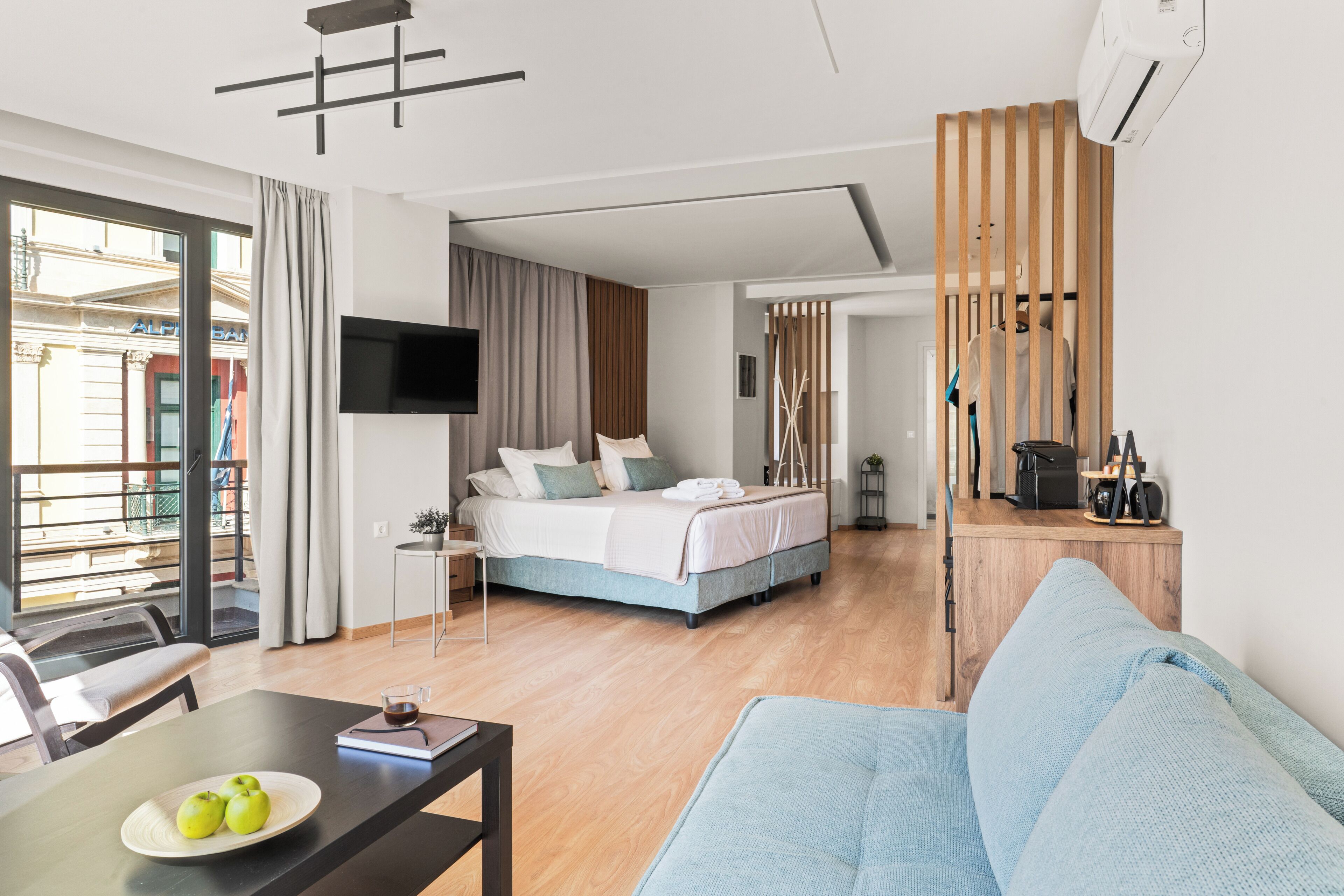 A modern hotel room with a bed, a TV, a dining area with a table and chairs, and a view of the outside.