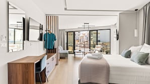 A modern hotel room with a wooden desk, a bed with a green headboard, a large window with a city view, and a balcony with outdoor seating. - Bedspot Heraklion (Heraklion)