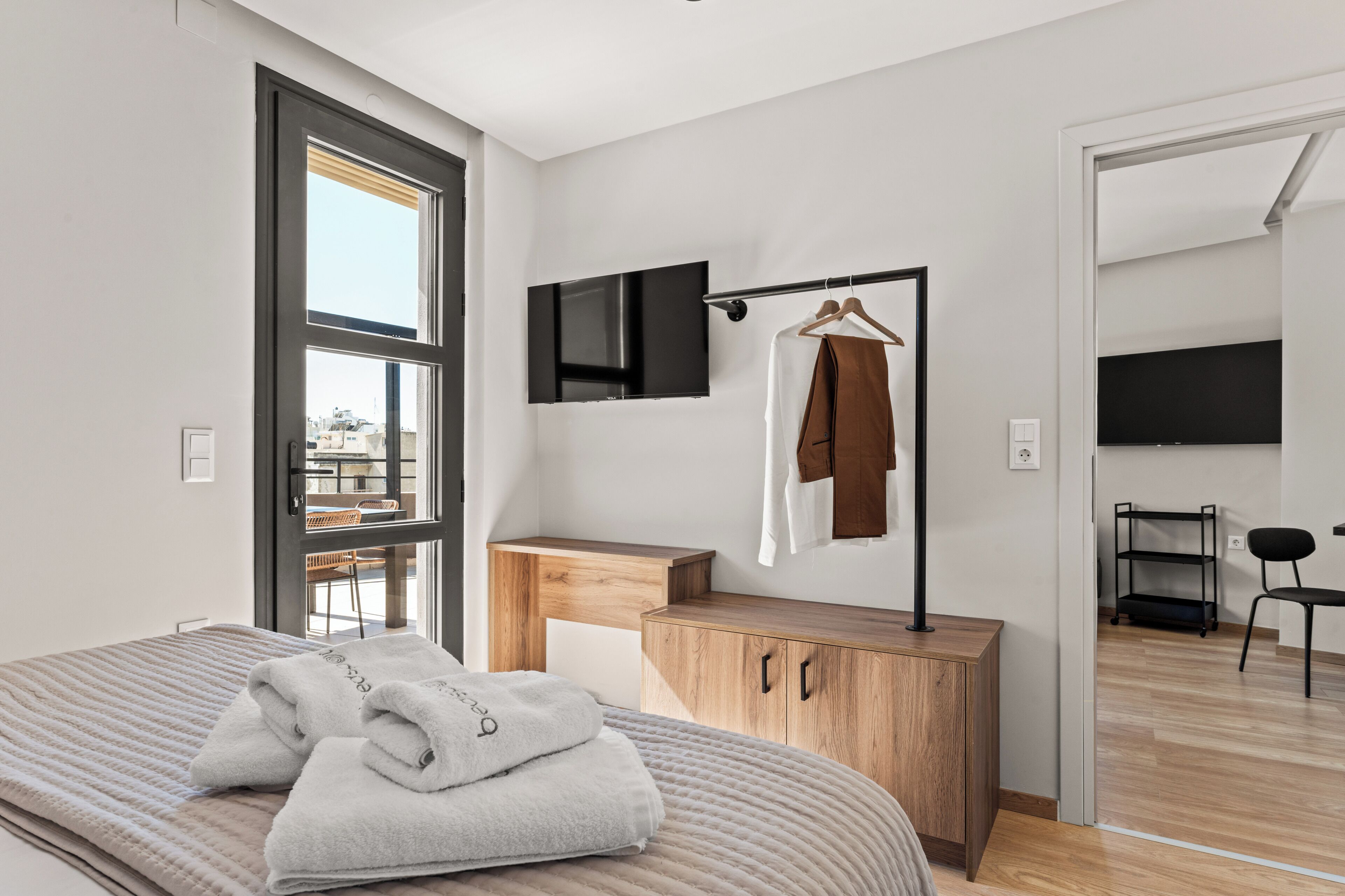 A modern bedroom with a bed, a wooden bedside table, a TV mounted on the wall, and a view of the outside through a large window.