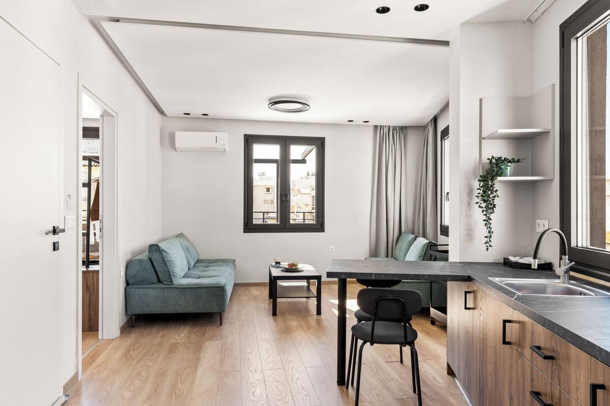 A modern kitchen with a central island, a dining area with a table and chairs, a sofa, and a view through a window.