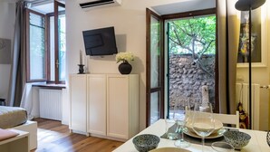 A modern room with a dining area, a TV mounted on the wall, and a view of a stone wall and trees outside. - Romantic loft with private garden | Self check-in (Verona)