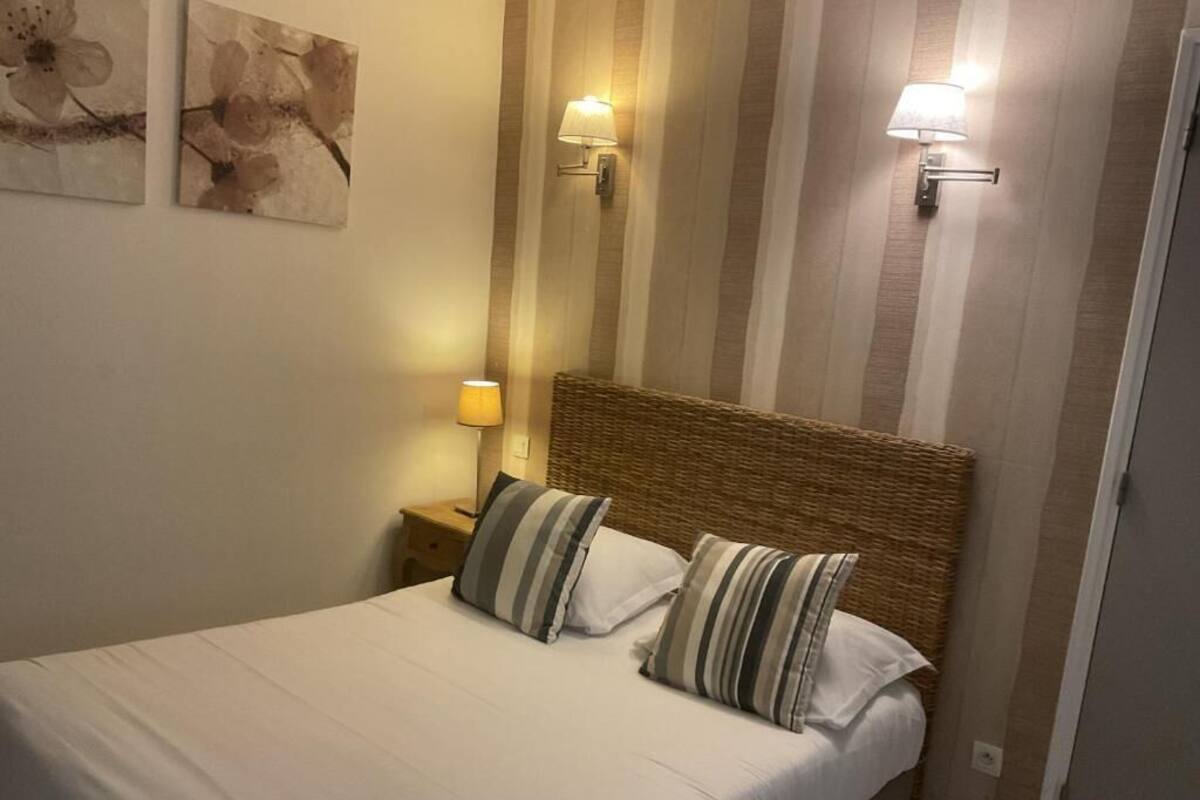 Un lit bien fait, agrémenté d’oreillers à rayures, d’une tête de lit en osier et de deux lampes fixées au mur.
