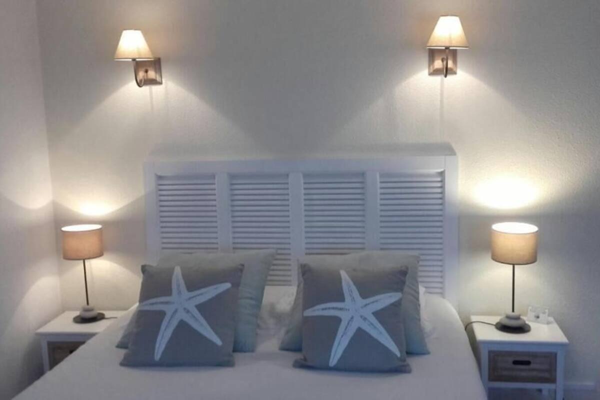 Une chambre à coucher comprenant un lit, deux tables de chevet, deux lampes fixées au mur et une fenêtre avec des volets.
