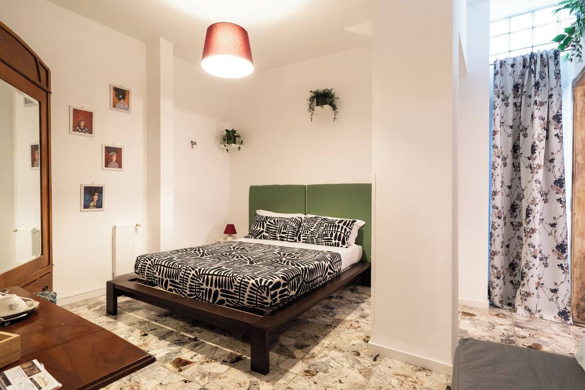 A bedroom with a bed, a wooden nightstand, a mirror, a red lamp, and a window with floral curtains.