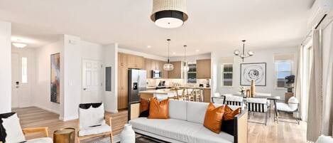 A modern living room with a sofa, coffee table, and chairs. There is a kitchen area with cabinets and a dining table. The room has a neutral color palette and a large window.