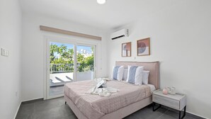 A bedroom with a bed, a nightstand, a window with a view of greenery, and an air conditioning unit.