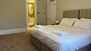 Uma cama bem arrumada com lençóis e toalhas brancas, uma mesa de cabeceira com abajur e uma porta que dá para o banheiro.