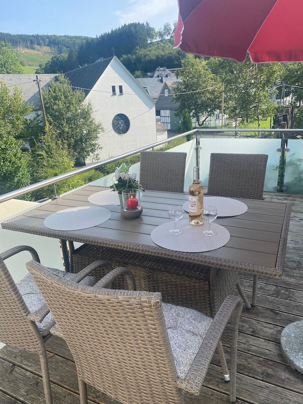Un área de comedor al aire libre con sillas de mimbre y una mesa lista para dos, con vistas a una iglesia y colinas boscosas.