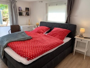 Un dormitorio con cama, una silla, una mesita de noche, una lámpara y una ventana con cortinas.