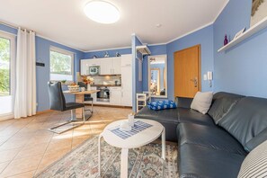 Un salon moderne avec un canapé, une petite table ronde, un coin cuisine doté de meubles blancs et une grande fenêtre avec des rideaux.