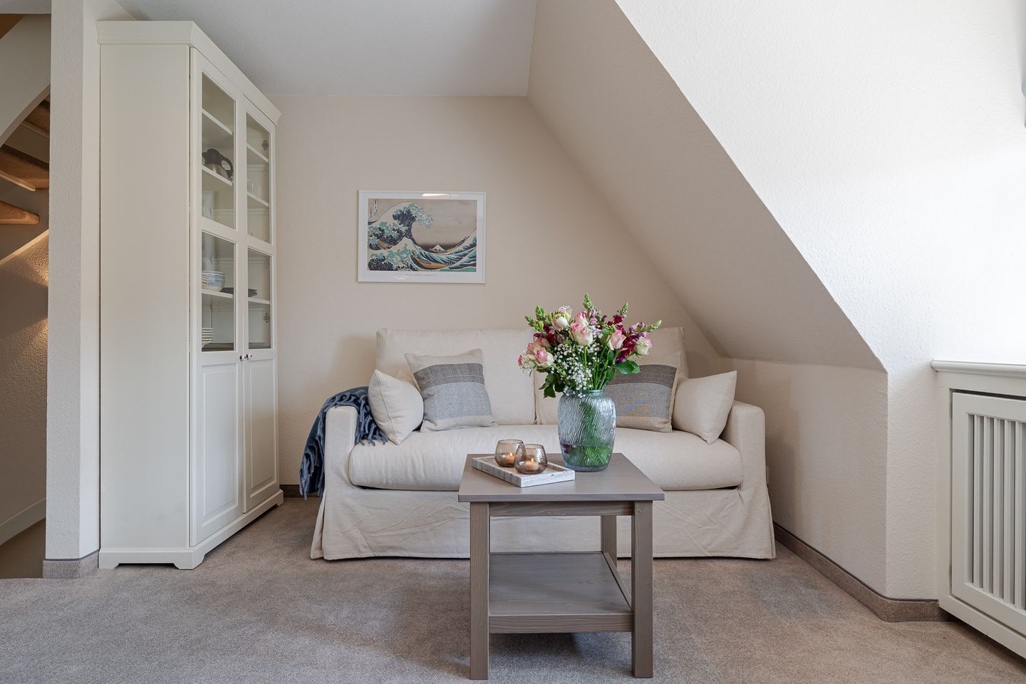 A cozy loft bedroom with a white sofa, a glass cabinet, a coffee table, and a framed artwork on the wall.