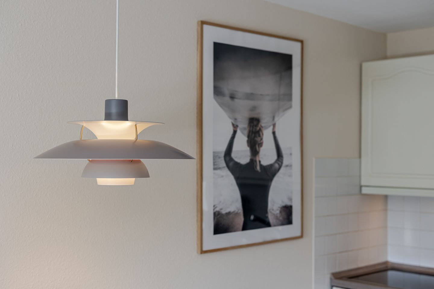 A pendant light fixture with a modern design, a framed photograph on the wall, and a kitchen area with white cabinets.