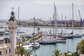 A marina with multiple sailboats docked, a prominent clock tower, and a large cargo ship in the background.