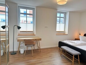 Un dormitorio moderno con suelo de madera, un escritorio, una silla, una cama y dos ventanas con persianas.