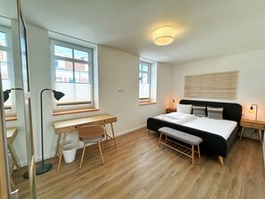 A bedroom with a bed, bedside tables, a desk, and a chair. There are windows with blinds, a ceiling light, and a wall-mounted shelf.