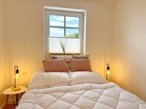 A bedroom with a bed, pillows, bedside lamps, a window with blinds, and a view of greenery outside. - Harbor master Björn - Spacious living above the rooftops of Sassnitz (Sassnitz)