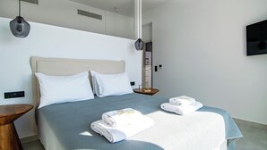 A modern hotel room with a large bed, two white pillows, a grey bedspread, and two towels on the bed. There is a wooden side table, a small round table with a book, and a wall-mounted TV. - 5-Bedroom Luxury Villas in Lefkada (Lefkada)