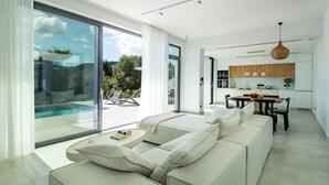 A modern living room with a large sliding glass door leading to a pool, a sofa set, and a dining area with a kitchen in the background. - 5-Bedroom Luxury Villas in Lefkada (Lefkada)