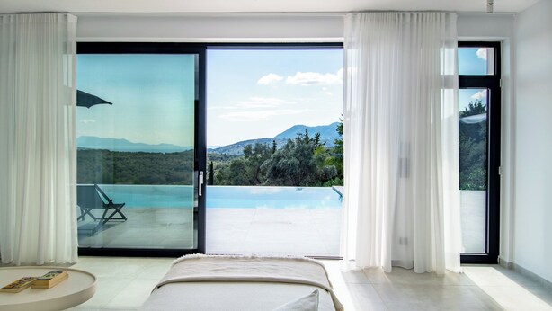 A modern bedroom with a bed, a small round table, and a view of a pool and mountains through large sliding glass doors. - 5-Bedroom Luxury Villas in Lefkada (Lefkada)
