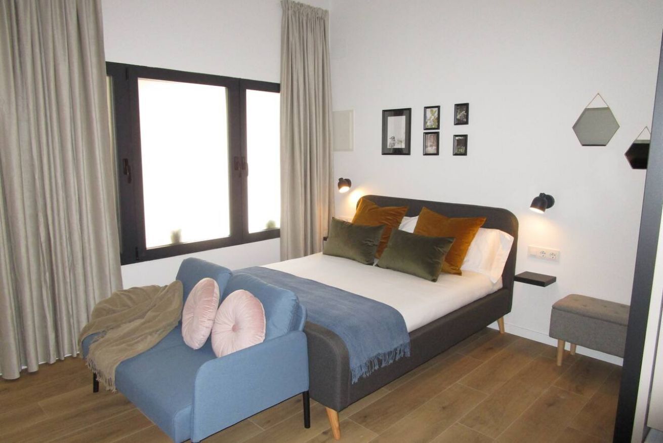 Um quarto moderno com uma cama, uma poltrona azul, uma mesinha de cabeceira e uma janela com cortinas.