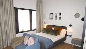 Un dormitorio moderno con cama, un sillón azul, una mesita de noche y una ventana con cortinas.