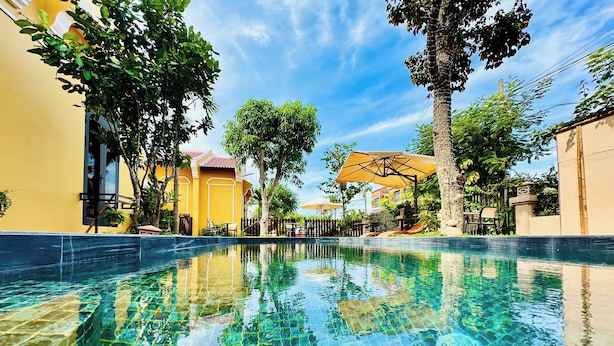 Piscina límpida com bordas azulejadas, cercada por vegetação exuberante e edifícios com fachadas amarelas e bege.