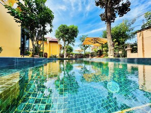 Una piscina limpia con bordes de azulejos, rodeada de vegetación exuberante y edificios con fachadas amarillas y beige.