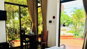 Quarto de hotel com cama, escrivaninha e cadeira. Tem uma janela com cortinas e vista para o verde lá fora.