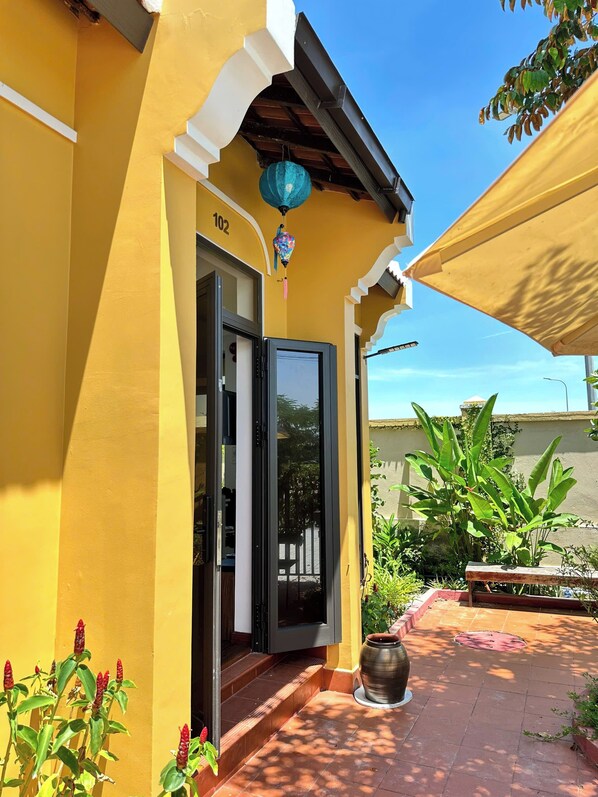 Un edificio amarillo con el número '102' en la puerta, una puerta negra y un patio con baldosas y plantas.
