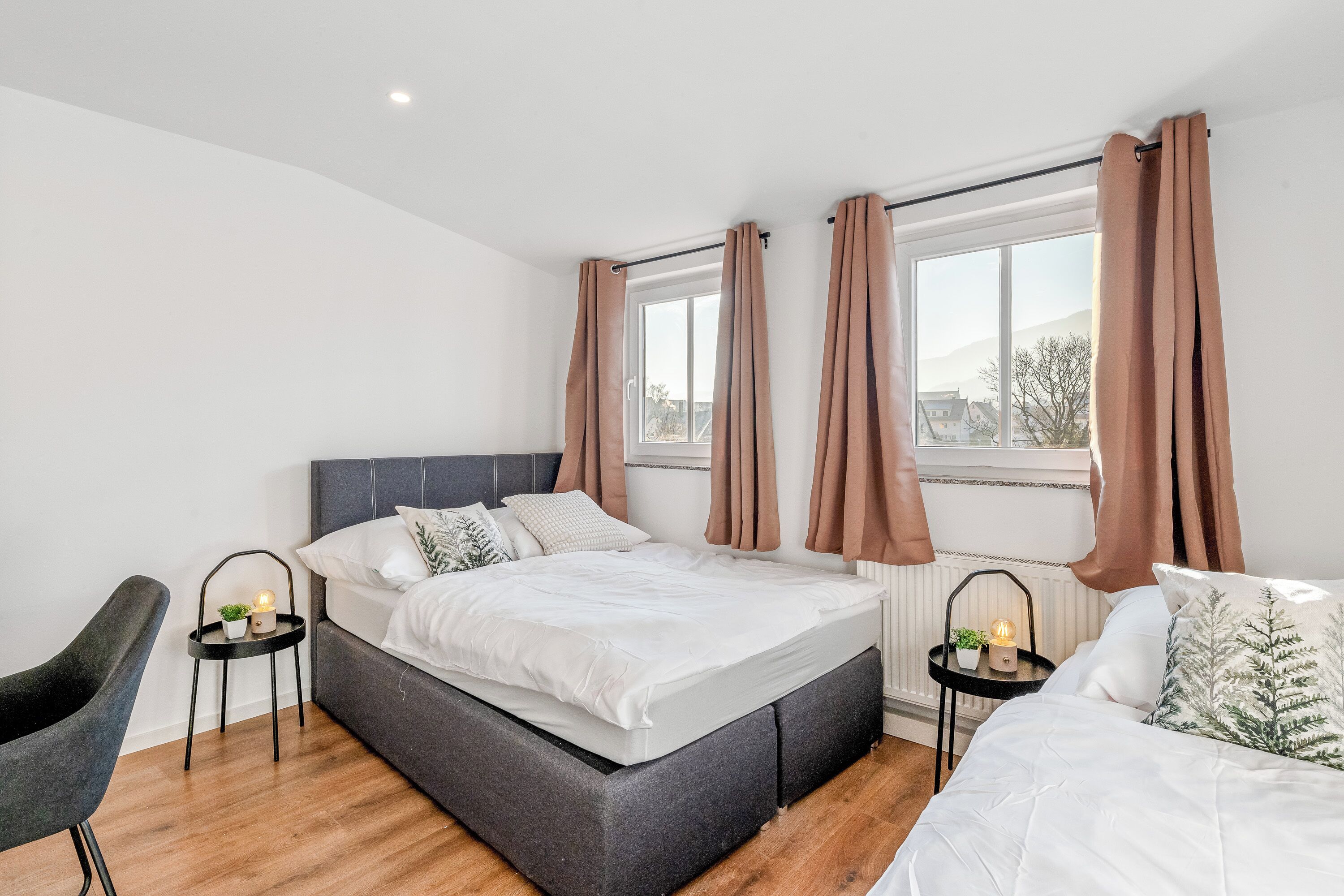 A modern bedroom with a large bed, two bedside tables with lamps, a chair, and a window with curtains.