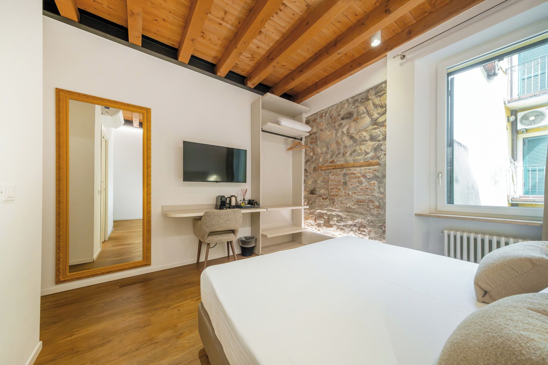 Ein Schlafzimmer mit Holzbalkendecke, Steinwand, Bett, Fernseher, Schreibtisch und Fenster.