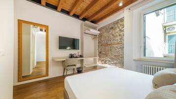Ein Schlafzimmer mit Holzbalkendecke, Steinwand, Bett, Fernseher, Schreibtisch und Fenster.