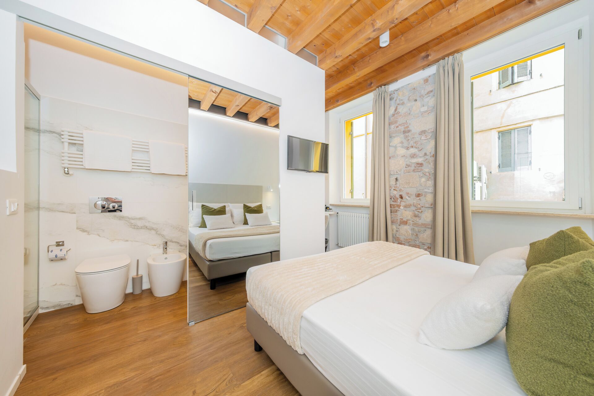 A modern bedroom with a wooden ceiling, a large bed with white bedding, a TV mounted above the bed, and a bathroom with a glass shower enclosure.